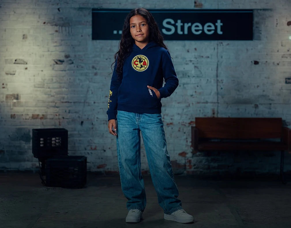 A young girl stands in a dimly lit warehouse wearing a navy blue Club América hoodie, blue jeans, and white sneakers. The background features a brick wall, and wooden bench.