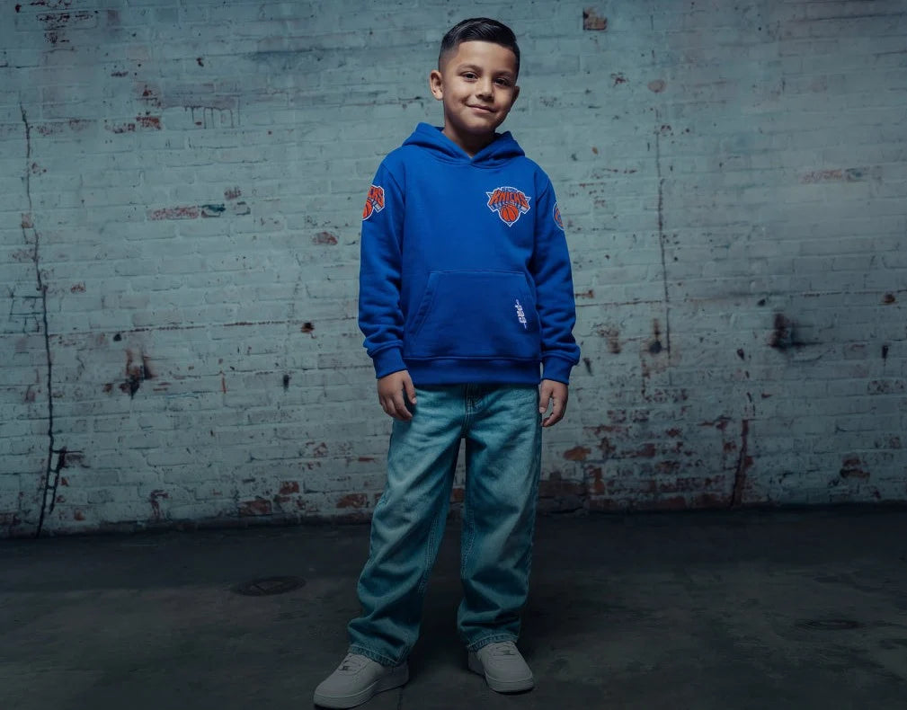 A young boy stands in front of a distressed white brick wall wearing a royal blue hoodie with a New York Knicks logo on the chest, paired with light blue jeans and gray sneakers.