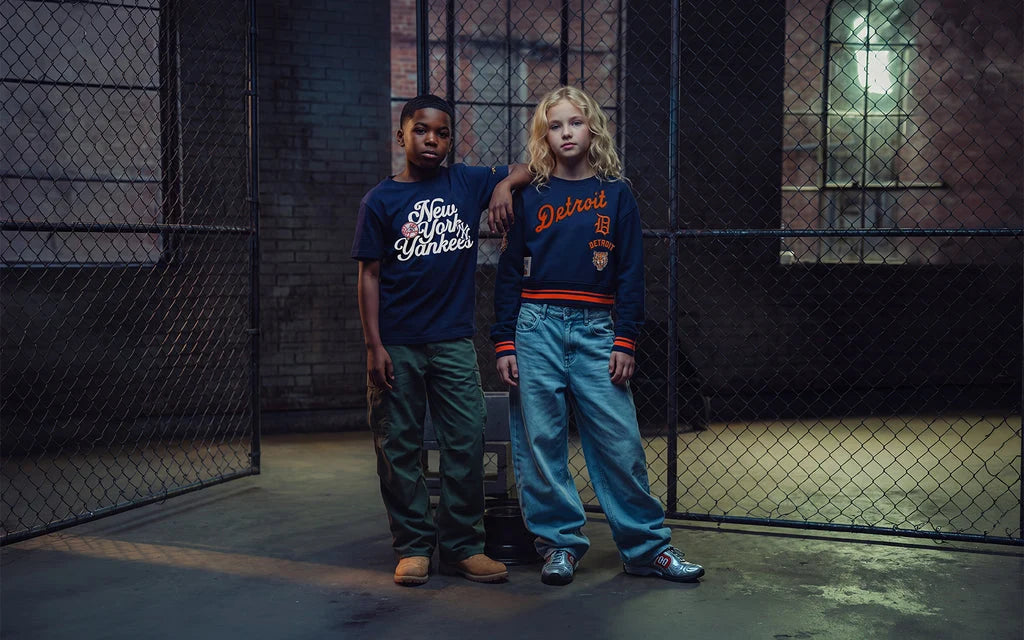 Two models standing in front of a chain-link fence in an industrial loft setting. The boy on the left wears a dark navy graphic t-shirt and green pants, while the girl on the right wears a dark navy Detroit Lions sweatshirt and light blue jeans.