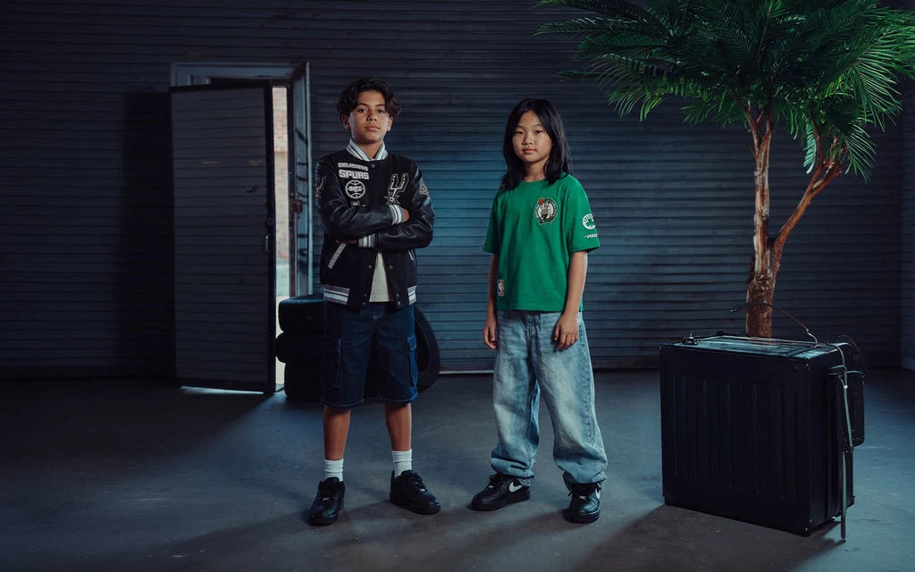 Two children modeling streetwear in a dark, moody studio setting. The child on the left wears a black varsity-style bomber jacket with white patches over a white shirt and dark shorts, while the child on the right wears a green crewneck t-shirt with a circular crest on the chest and light-wash jeans.