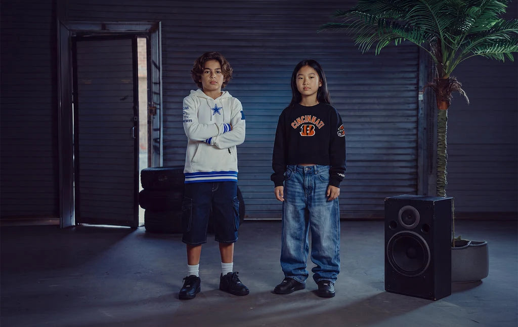 Two children standing in a dark industrial room with a garage door background, a potted palm, and a large speaker. The child on the left wears a white hoodie with blue trim and a star logo, and the child on the right wears a black sweatshirt with orange lettering and blue jeans.