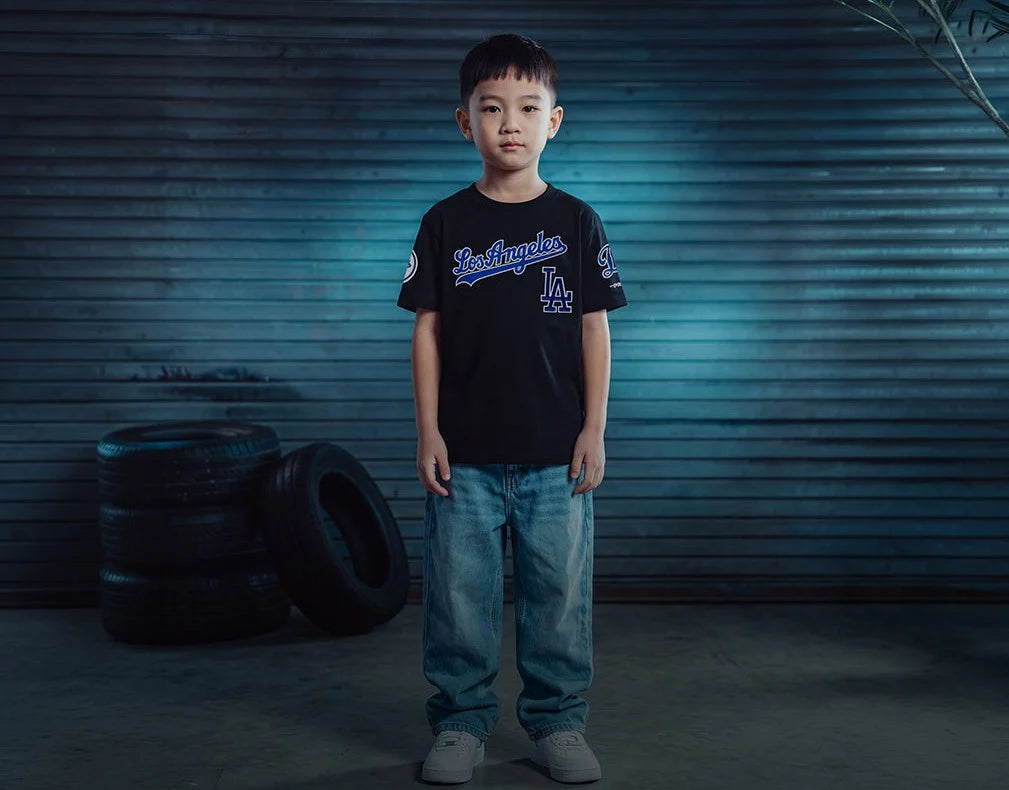 A young boy stands in front of a blue-toned corrugated metal wall, wearing a black short-sleeve graphic T-shirt with blue and white lettering, blue jeans, and gray sneakers. Two black tires are stacked on the floor to his left.