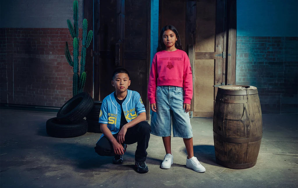 Two children modeling streetwear in a moody industrial loft setting. The boy crouches wearing a light blue graphic t-shirt and dark jeans, while the girl stands wearing a bright pink cropped sweatshirt, denim shorts, and white sneakers.
