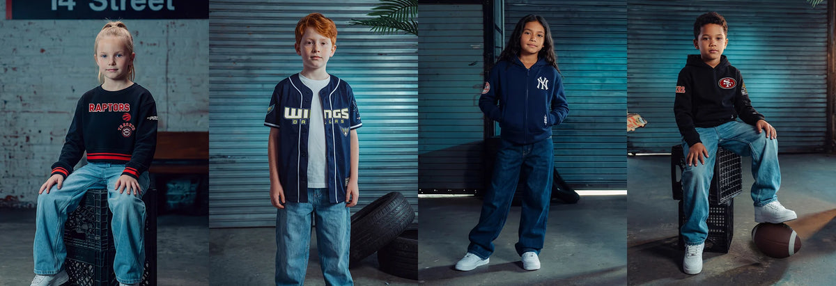 Four children modeling different streetwear outfits in a dark, industrial warehouse setting with corrugated metal walls. The outfits feature navy, black, white, and blue tones with various graphic details.